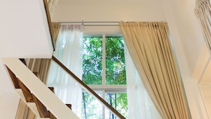 Un escalier avec une rampe en bois, une balustrade en verre et une vue sur la verdure à l’extérieur, visible à travers une fenêtre ornée de rideaux beiges.