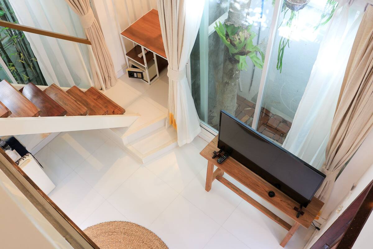A modern living room with a wooden staircase, a flat-screen TV on a wooden stand, and a glass door leading to an outdoor area with plants.