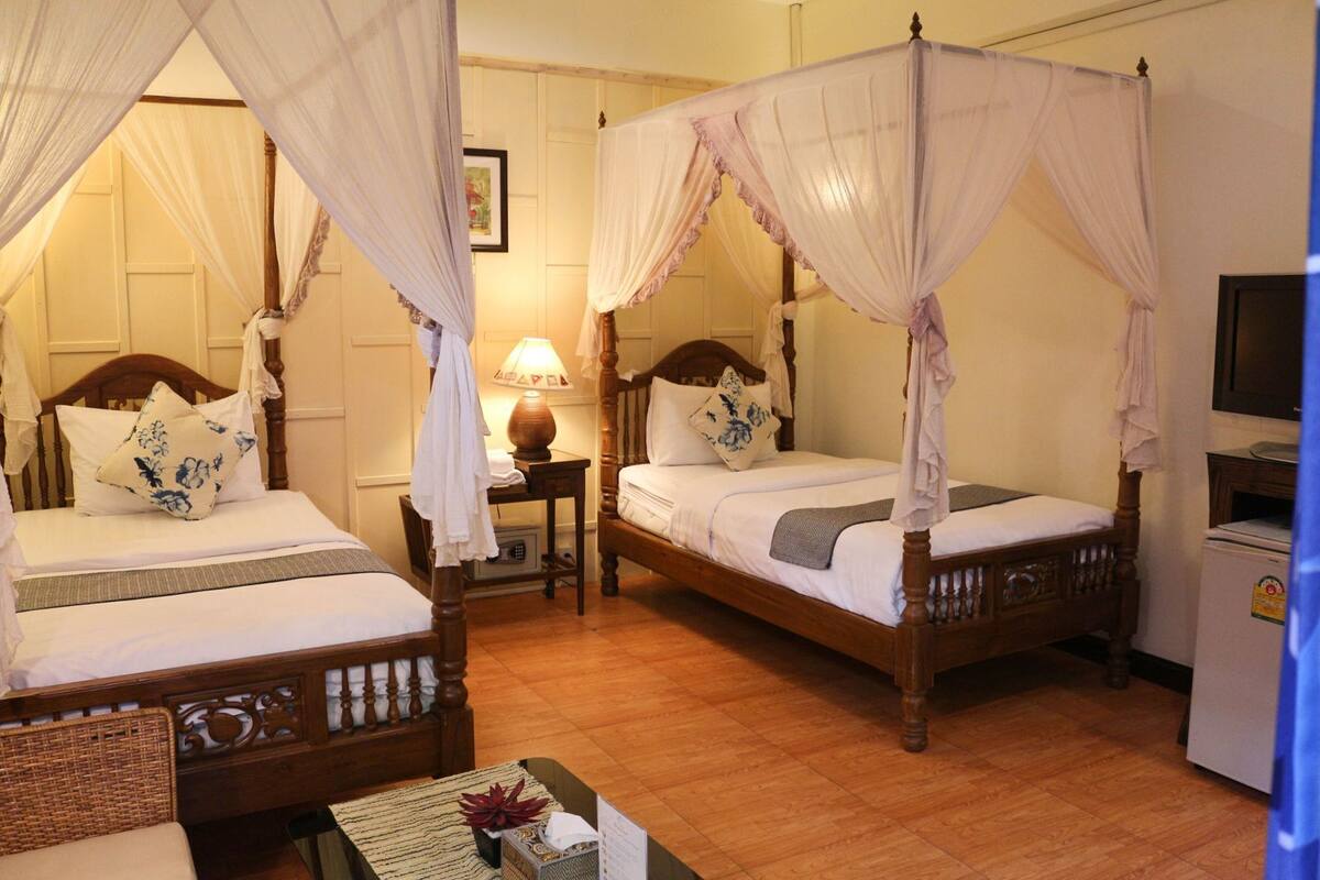 A room with two four-poster beds, a small table with a lamp, and a refrigerator.