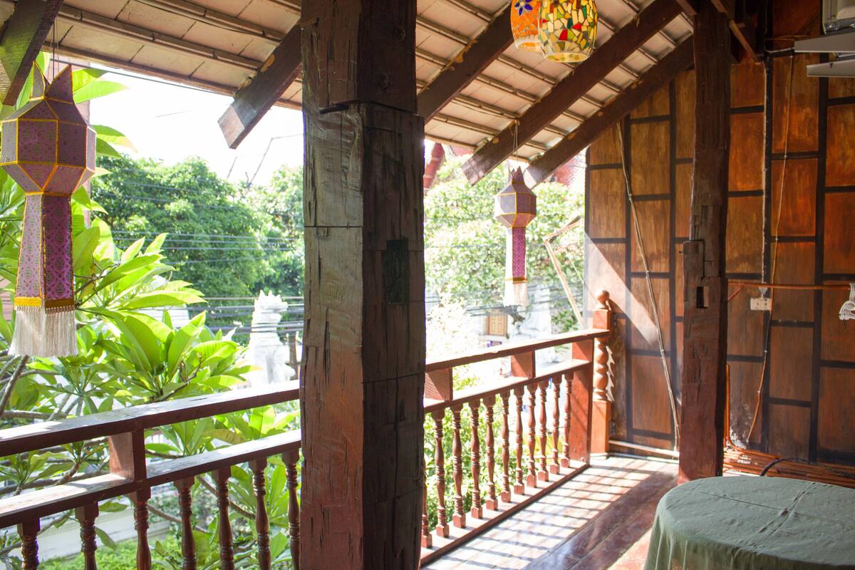 A wooden balcony with a railing, a tiled floor, and a view of greenery and buildings in the background.