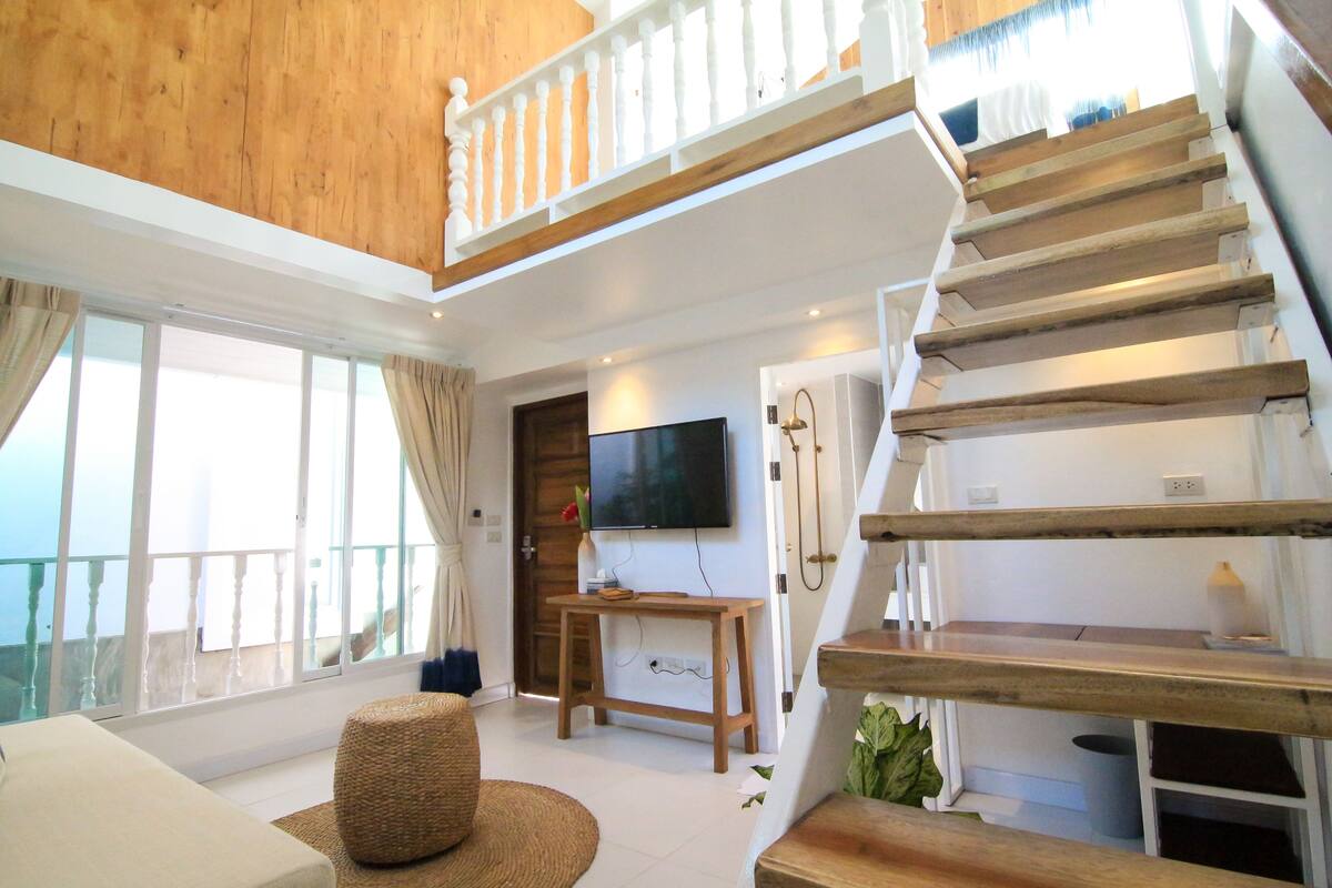 A spacious living area with a wooden staircase, a flat-screen TV, and large windows offering a view of the outdoors.
