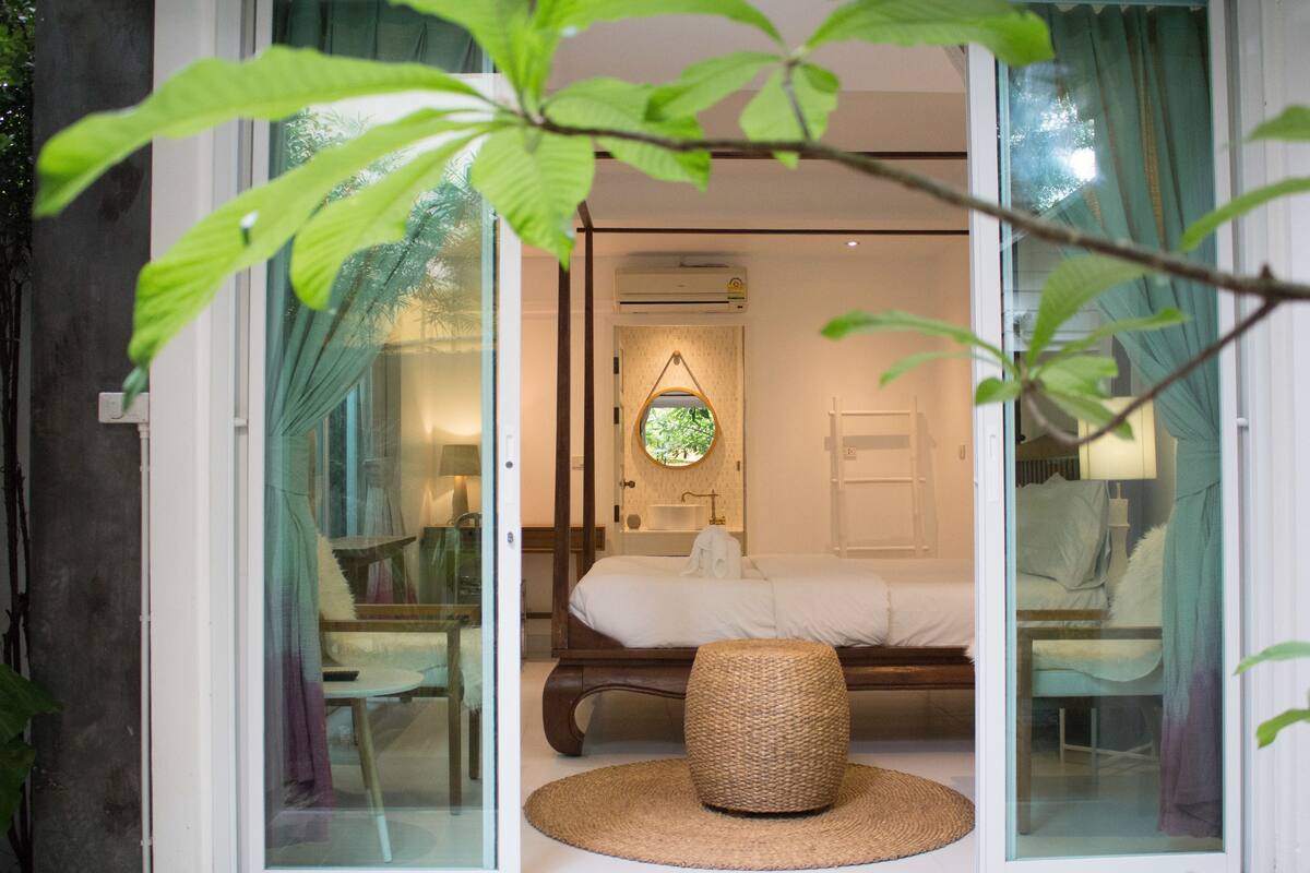 A bedroom with a bed, a round woven stool, and a circular mirror on the wall.