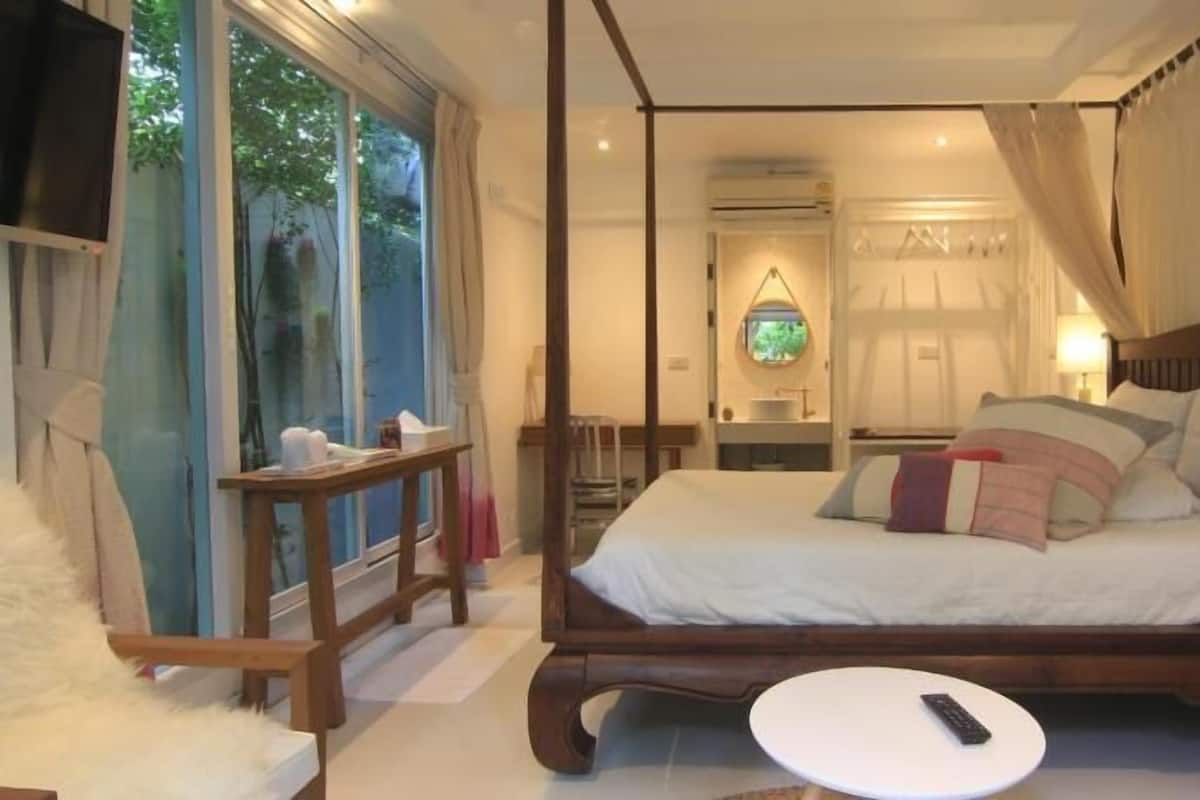 A four-poster bed with a canopy, a bedside table, a chair, and a view of the outdoors.