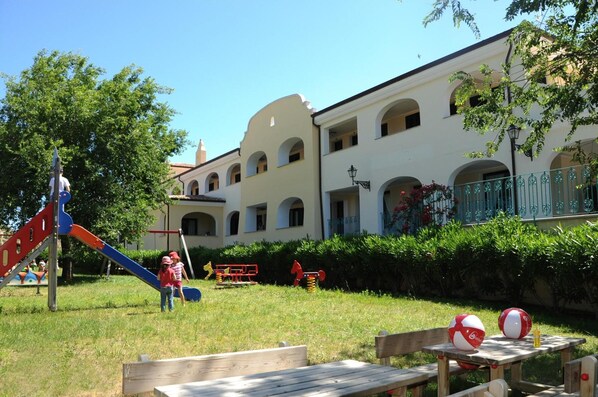 A playground with a slide and swings, a picnic table with a ball, and a building with arched windows and balconies. - Residence Cala Liberotto (Orosei)