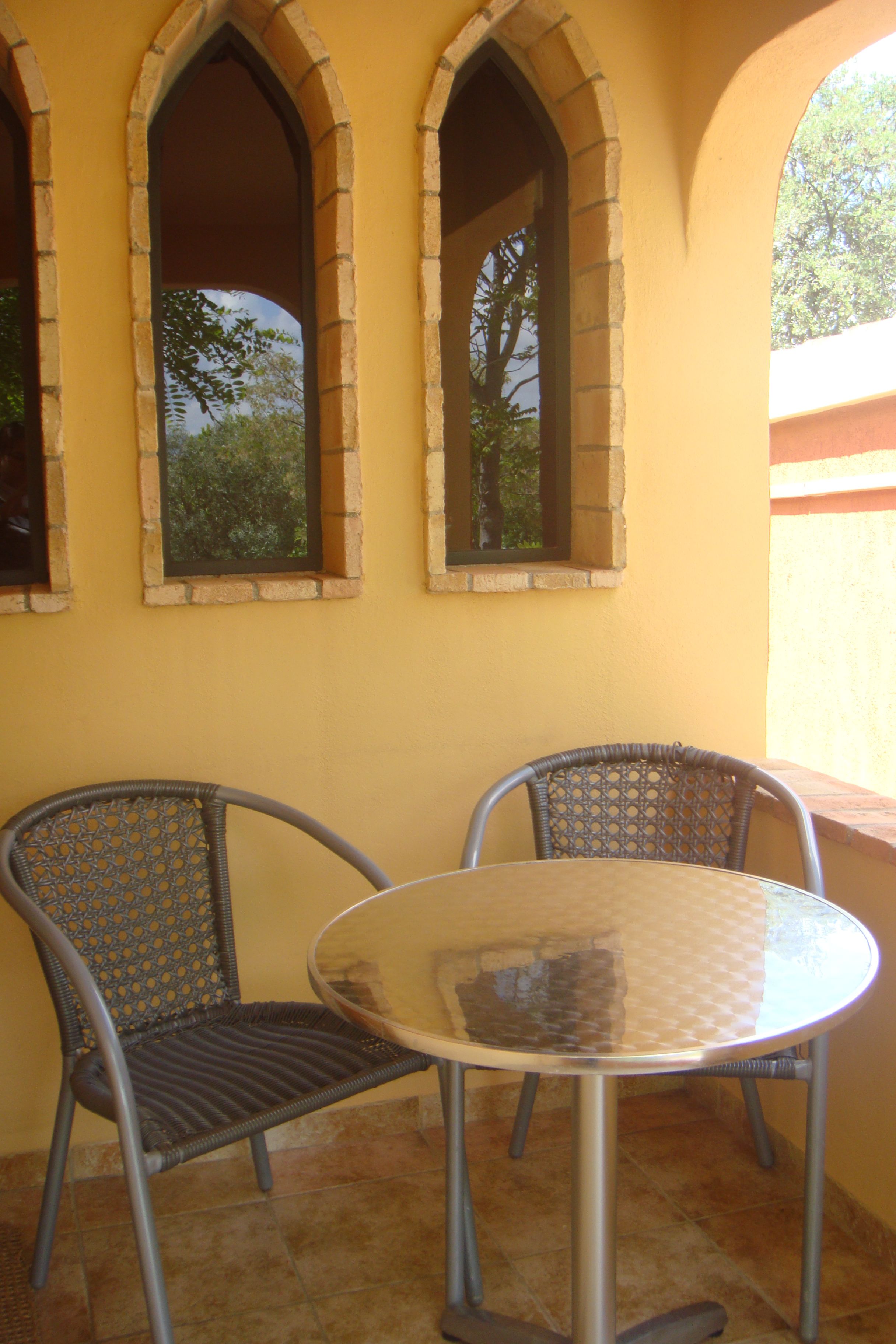 A patio with a round table and two wicker chairs, set against a yellow wall with three arched windows.
