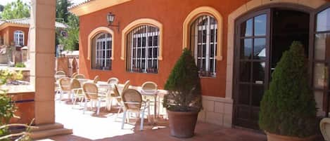 An outdoor patio with tables and chairs, a building with arched windows and a door, and potted plants.