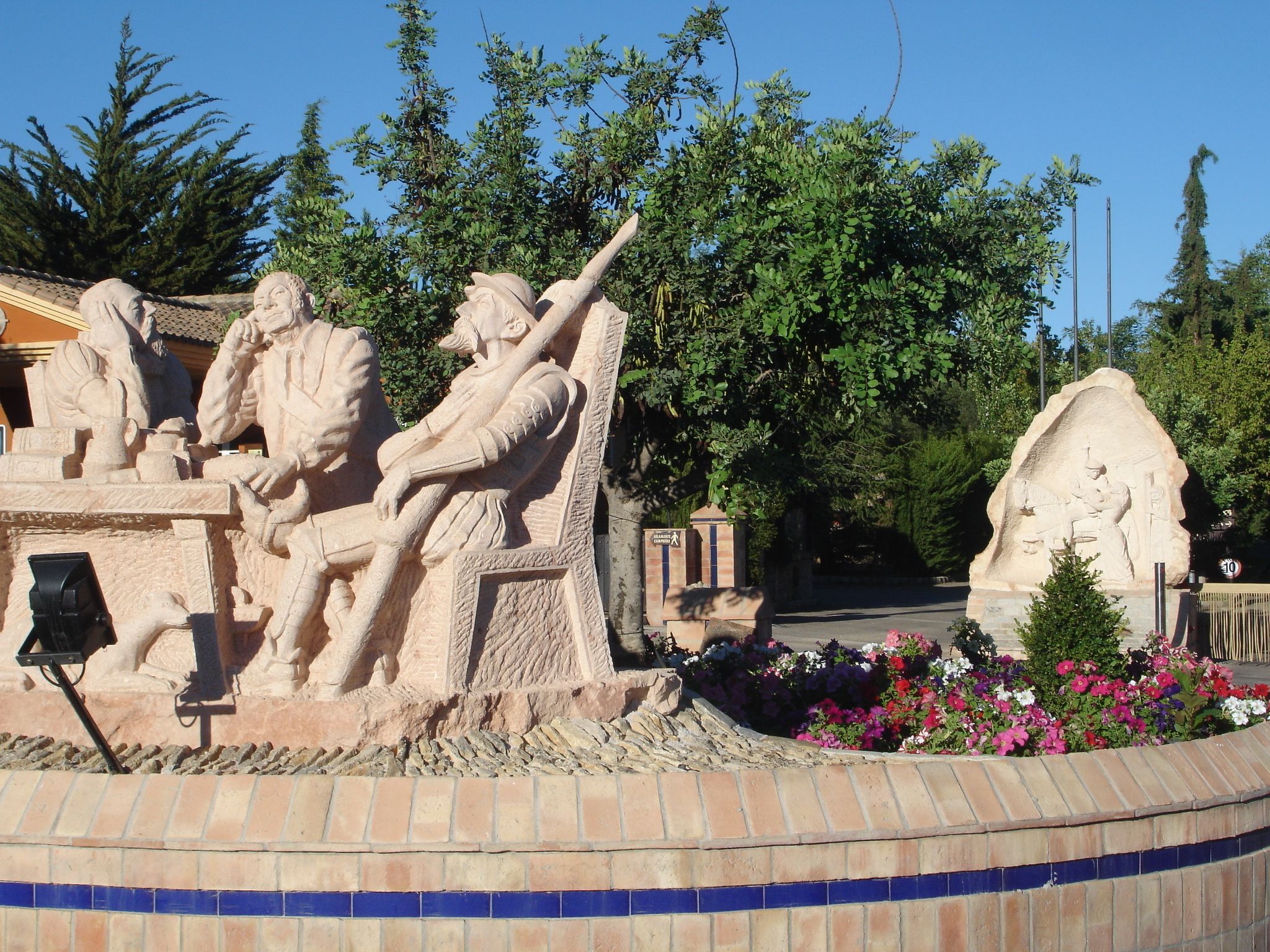 Une sculpture en pierre représentant une figure assise tenant un bâton, entourée d’une bordure en briques et de fleurs aux couleurs vives.
