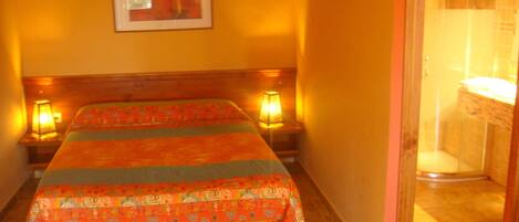 A bedroom with a bed, two bedside lamps, a framed picture on the wall, and a bathroom visible through an open door.