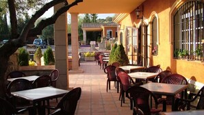 Ein Straßencafé mit Tischen, Stühlen und einer überdachten Sitzgelegenheit, mit Blick auf eine Straße mit parkenden Autos und Grünflächen.