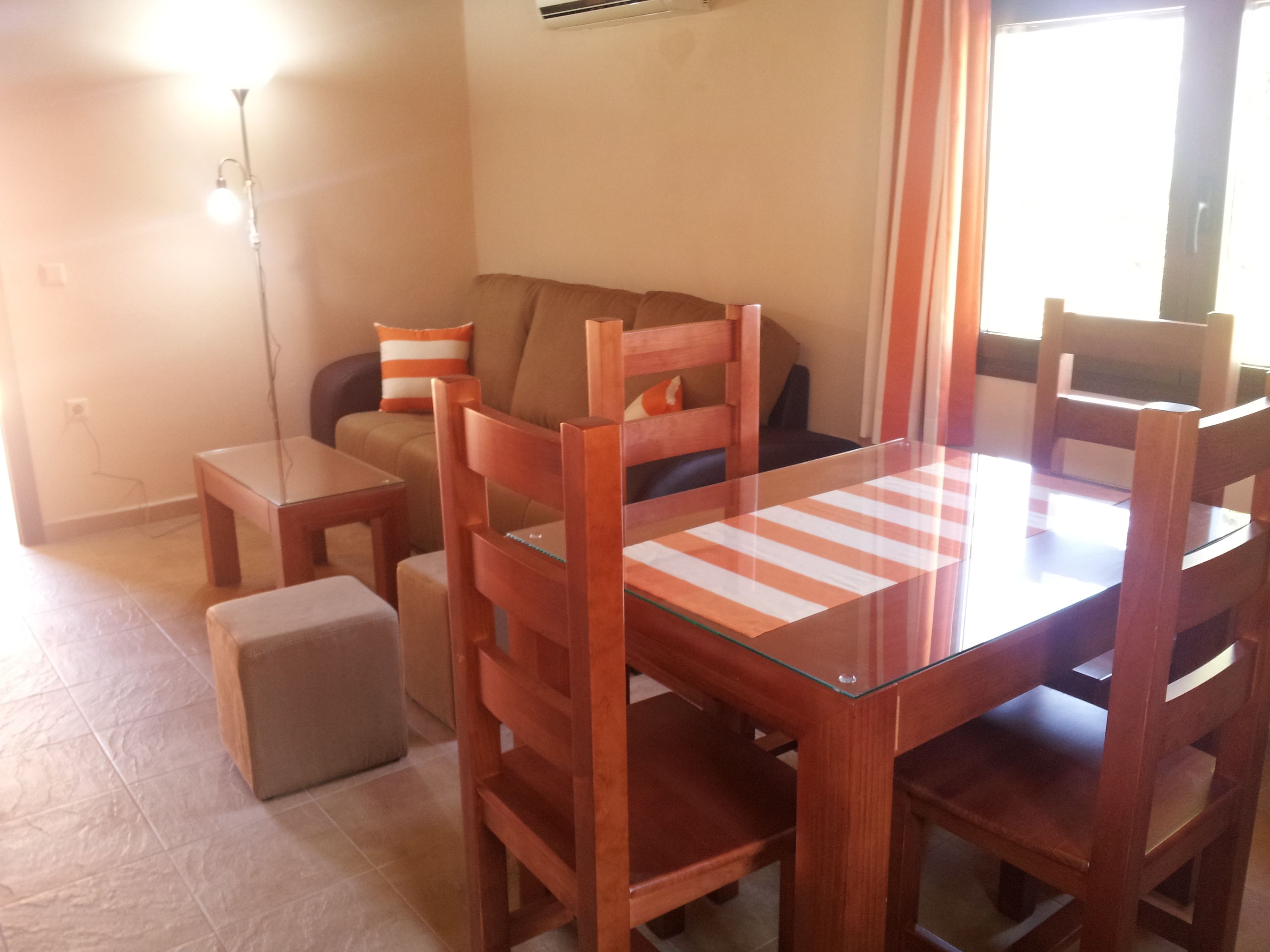A small dining area with a glass-top table and wooden chairs. There is a sofa with cushions in the background.