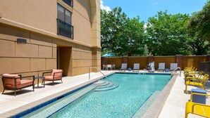 A rectangular pool with a clear blue edge, surrounded by yellow and orange lounge chairs, and a brick wall with a window and a door. - Hampton Inn & Suites Pensacola/Gulf Breeze (Gulf Breeze)