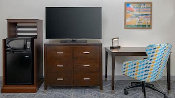 Habitación de hotel con televisión de pantalla plana, microondas, refrigerador pequeño, escritorio de madera, silla de oficina con diseño y una obra de arte abstracta enmarcada en la pared.