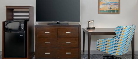 Habitación de hotel con televisión de pantalla plana, microondas, refrigerador pequeño, escritorio de madera, silla de oficina con diseño y una obra de arte abstracta enmarcada en la pared.
