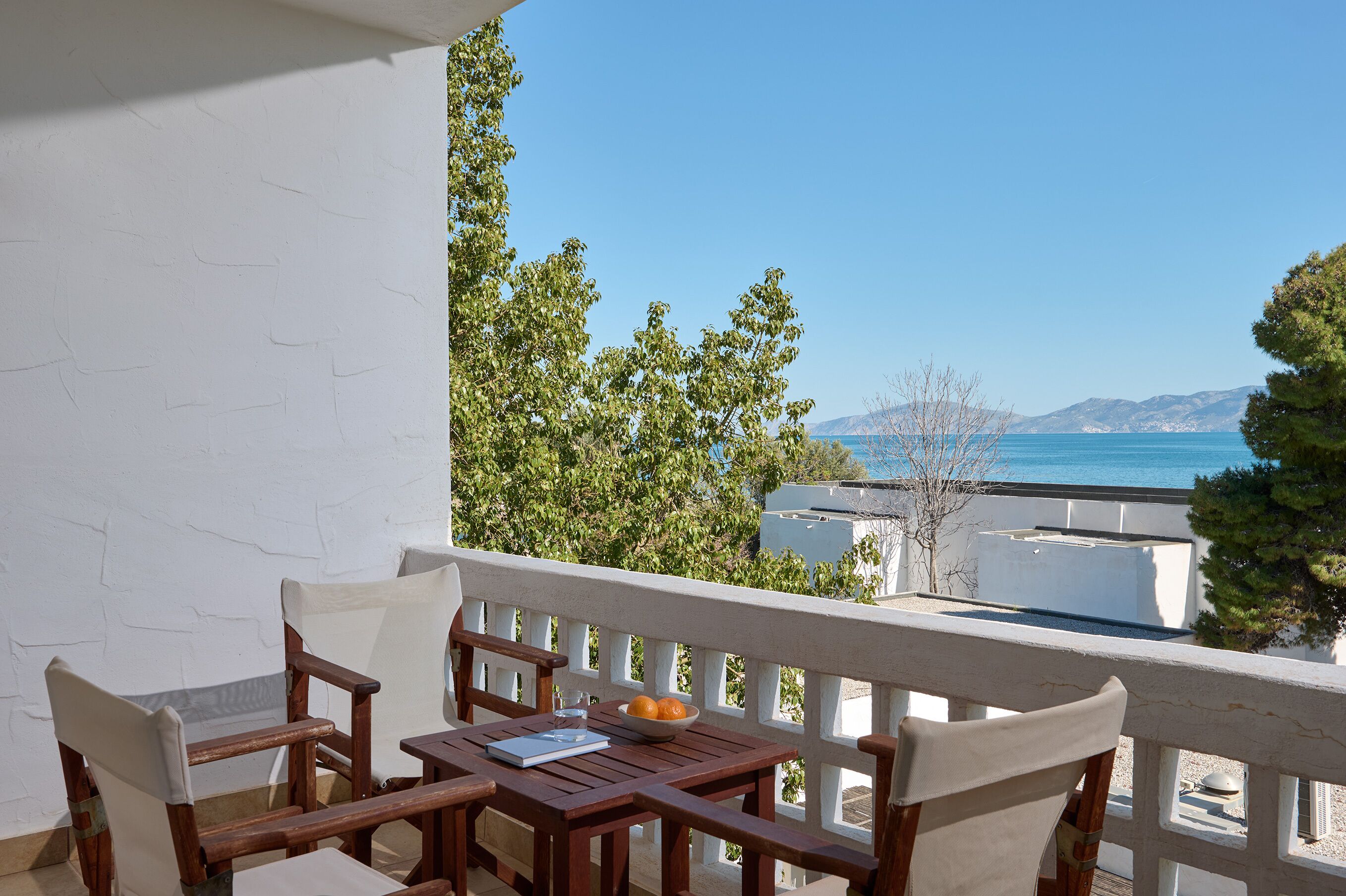 A balcony with a wooden table and chairs, a bowl of fruit, and a clear view of the sea and mountains.