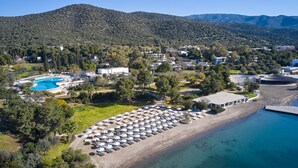 Vista aérea de un complejo turístico costero con piscina, sombrillas de playa y montañas al fondo.