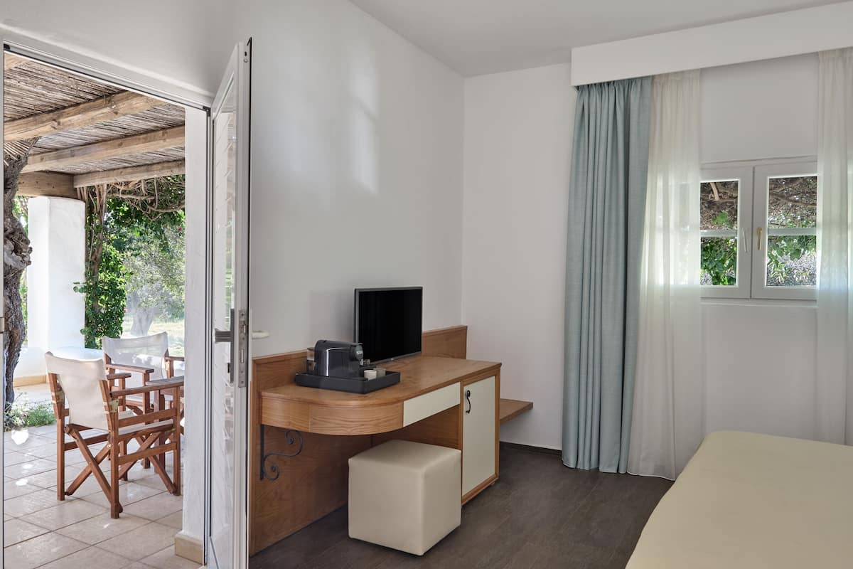 A hotel room with a wooden desk, a television, a bed, and a view of greenery outside.