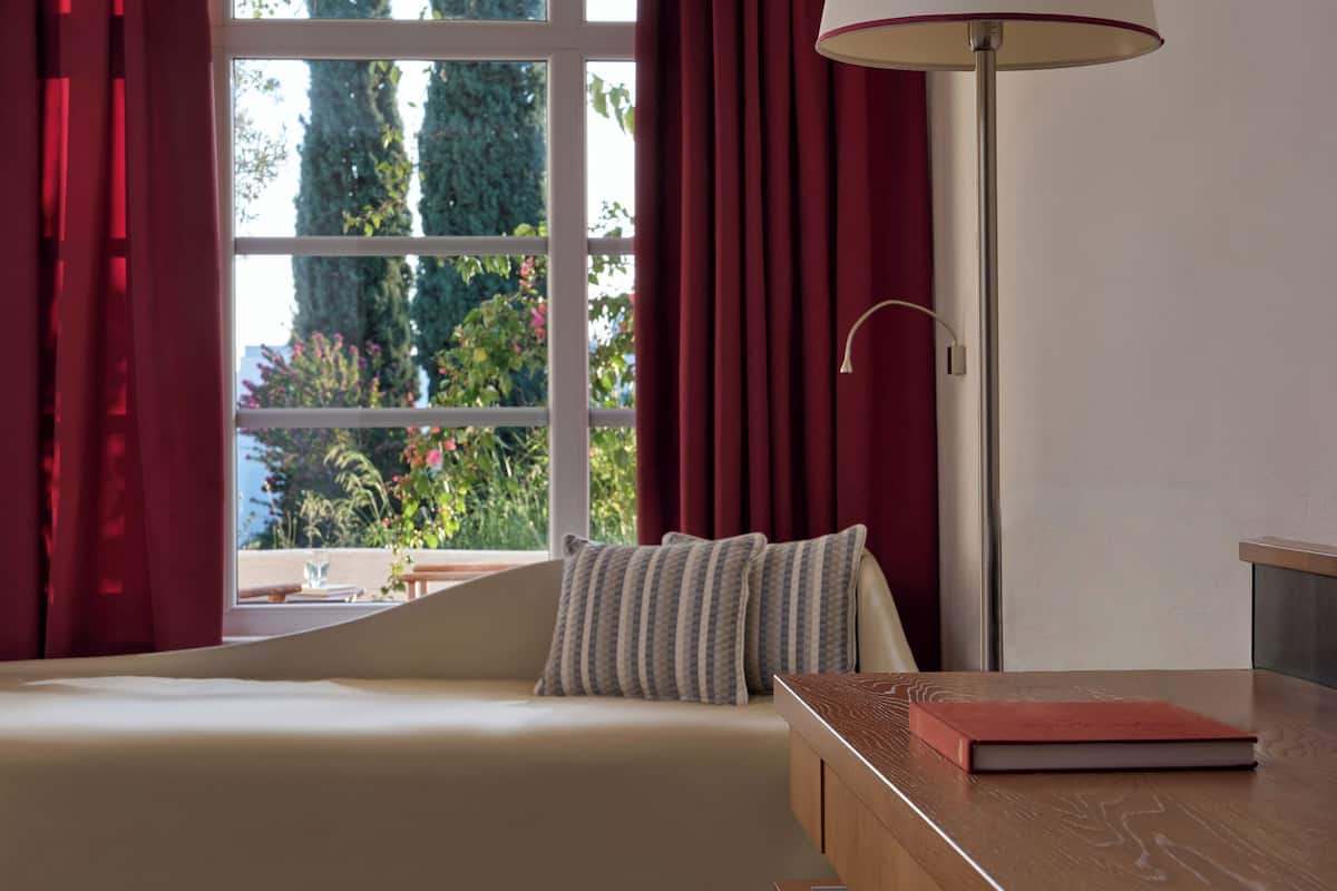 A room with a bed, a bedside table with a book, a floor lamp, and a window with curtains.