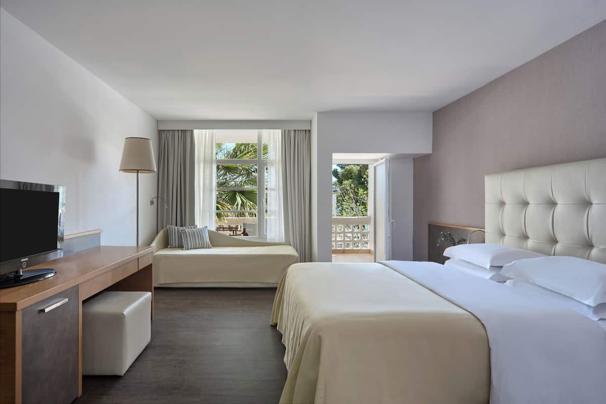 A hotel room with a bed, a desk with a TV, a window with curtains, and a balcony.