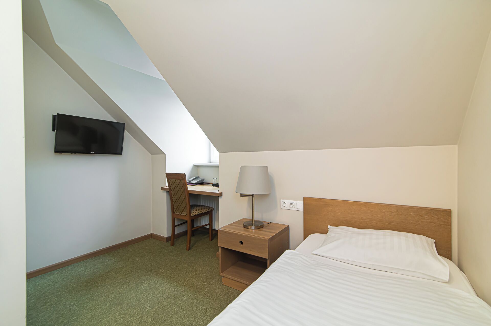 Una camera d'albergo con un letto, una scrivania con una sedia, una lampada e una TV fissata al muro.