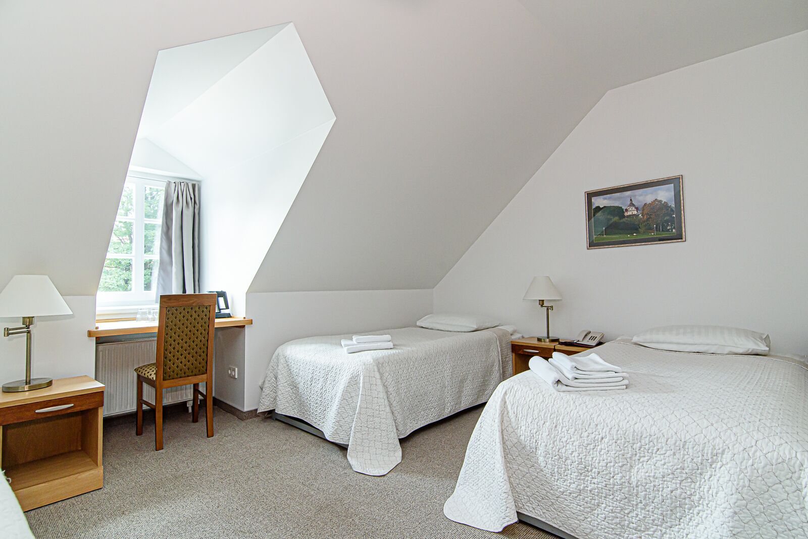 A hotel room with two beds, a desk, a chair, a window with curtains, a bedside table with a lamp, and a framed picture on the wall.
