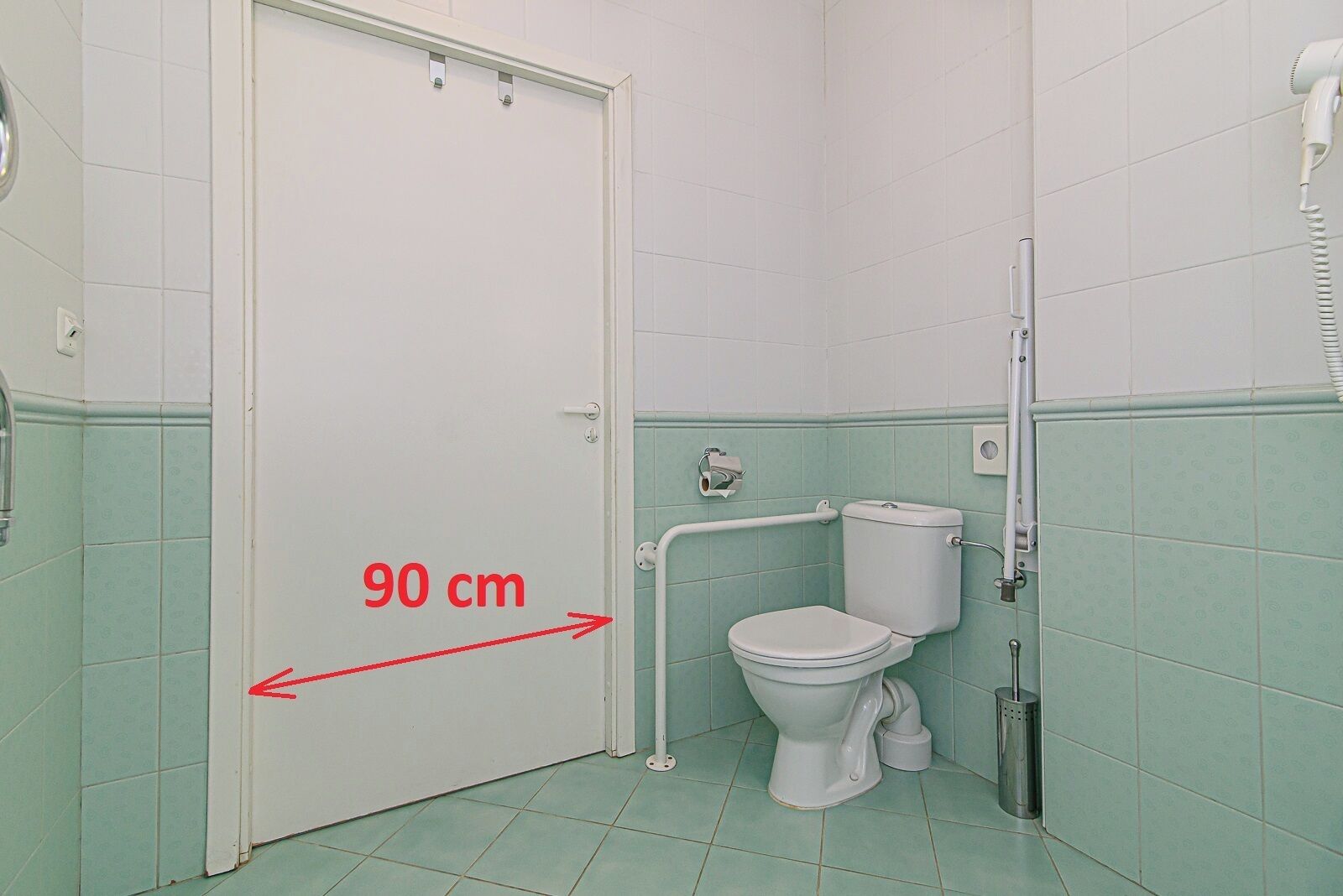 A bathroom with a white toilet, a handrail, and a door marked with a 90 cm measurement.