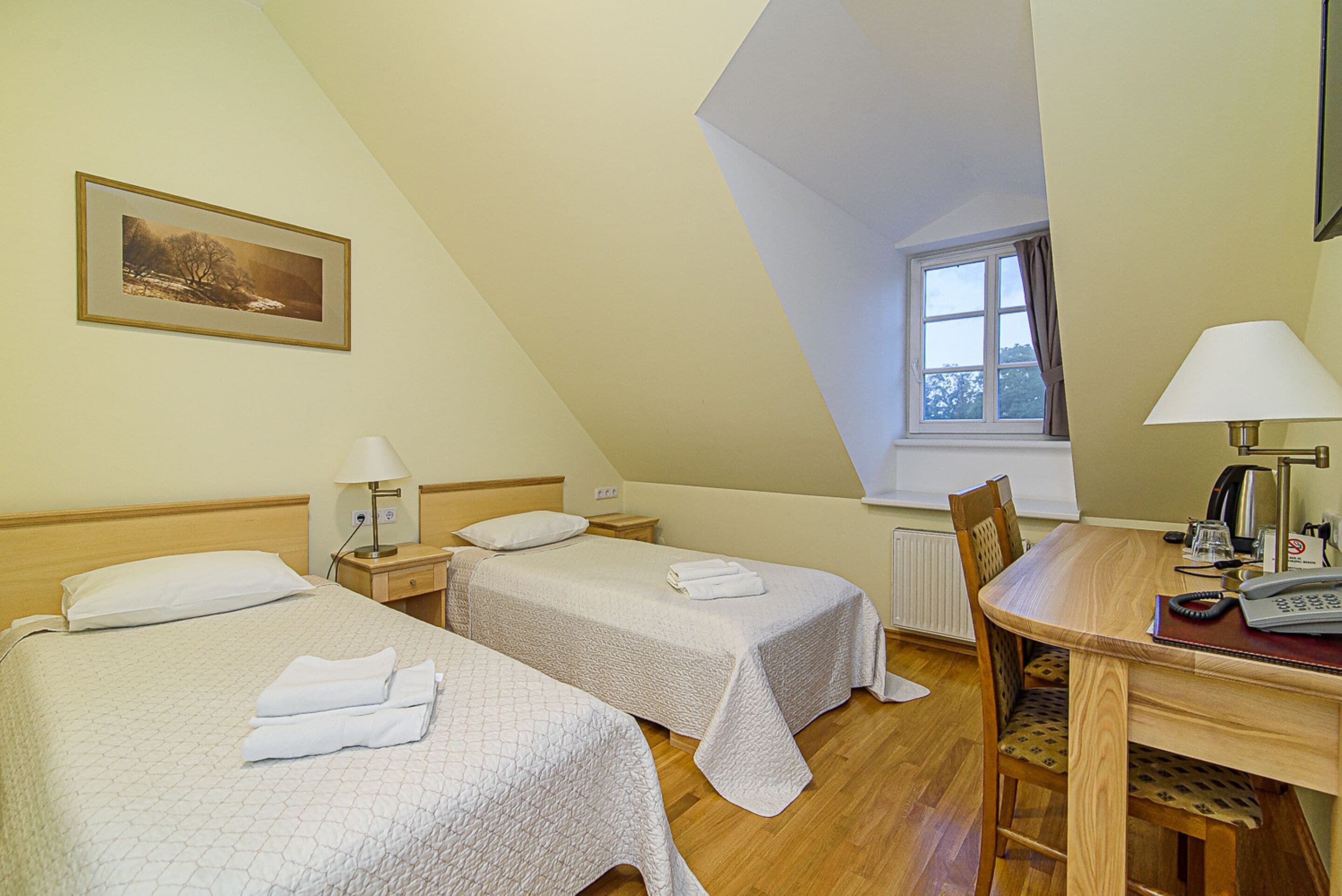 A room with two beds, a wooden table, a chair, a window, and a painting on the wall.