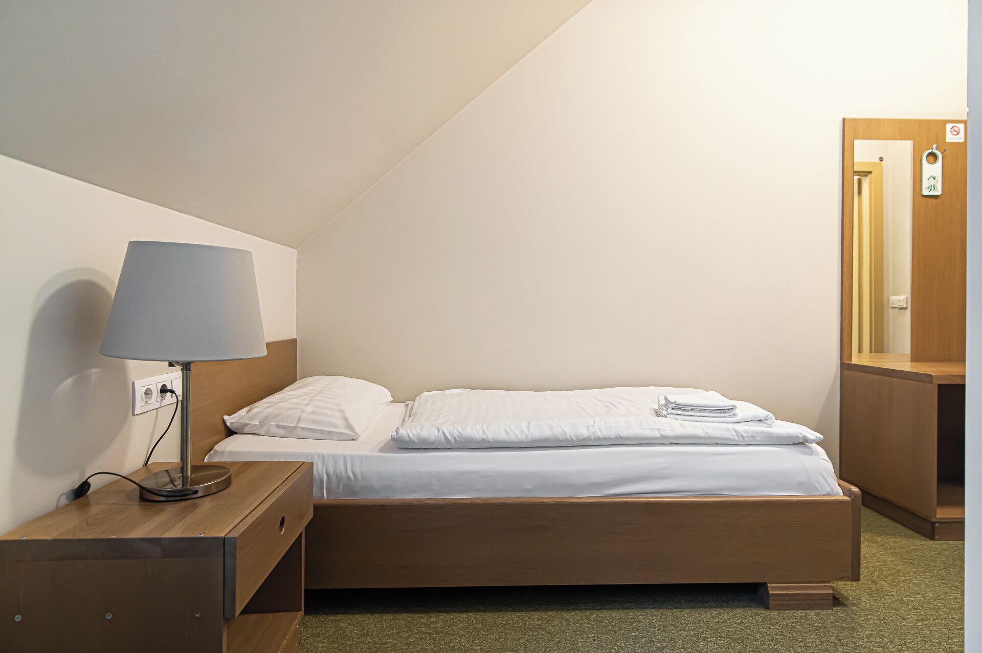 A single bed with white linens, a bedside table with a lamp, and a wooden nightstand.