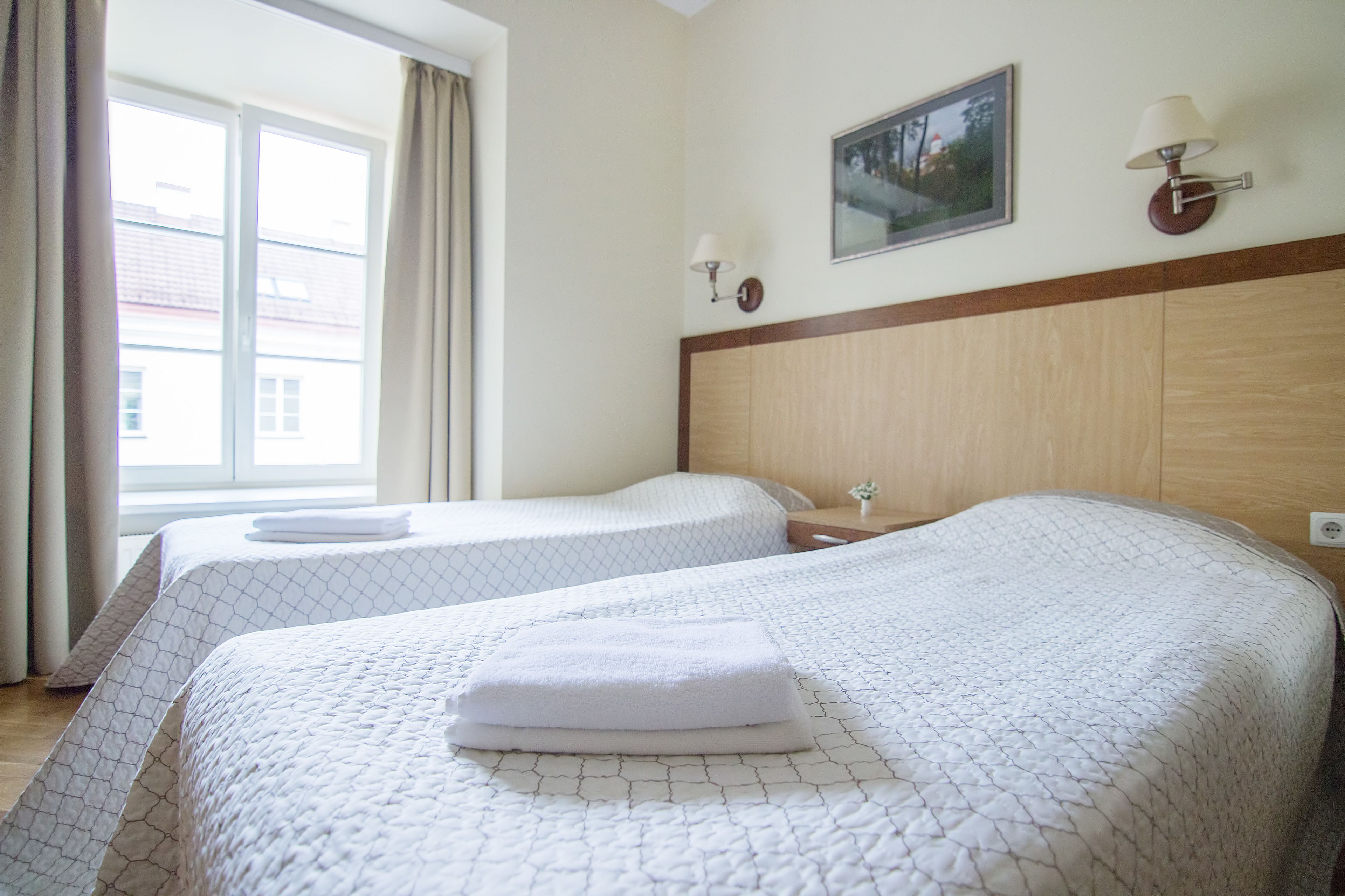 Habitación de hotel con dos camas, una ventana con cortinas, una lámpara de pared y un cuadro enmarcado.