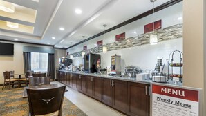 A hotel kitchen area with a weekly menu sign, wooden cabinets, and a dining area with chairs and a table. - Best Western Plus Longhorn Inn & Suites (Luling)