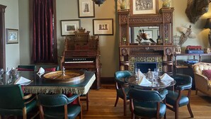 Un comedor con piano, sillas tapizadas de verde, una araña, obras de arte enmarcadas y una cabeza de animal montada.
