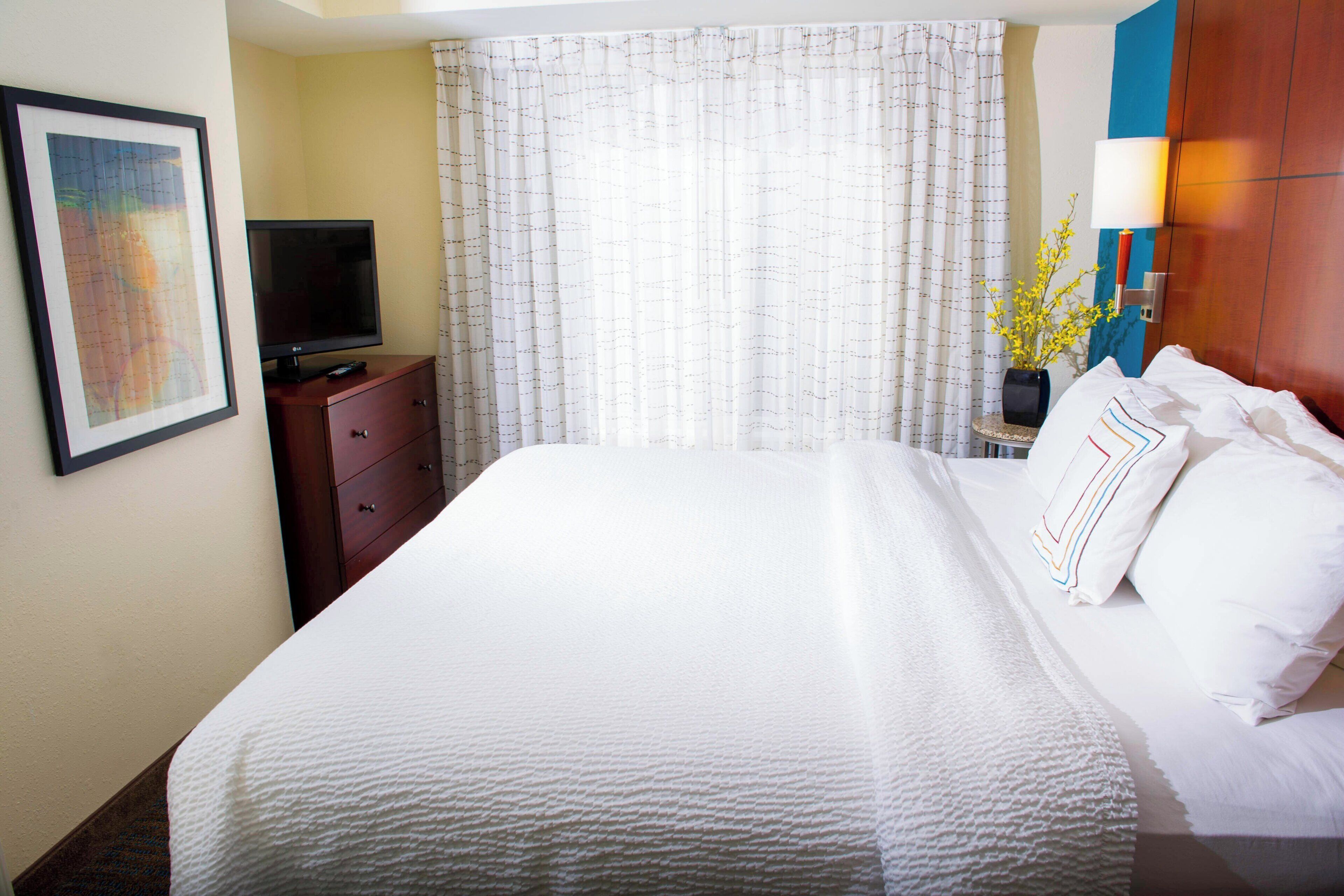A hotel room with a large bed, a television on a wooden dresser, and a window with curtains.