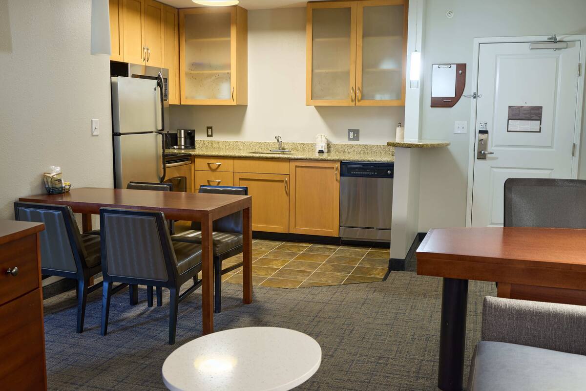 A compact kitchen with wooden cabinets, a stainless steel refrigerator, a dishwasher, and a dining area with a table and chairs.