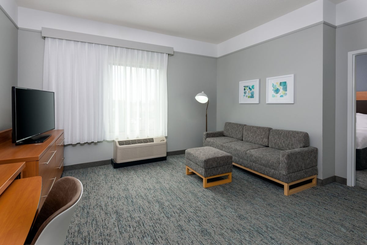 A hotel room with a flat-screen TV, a grey sofa, a small ottoman, a desk with a chair, and a standing lamp.