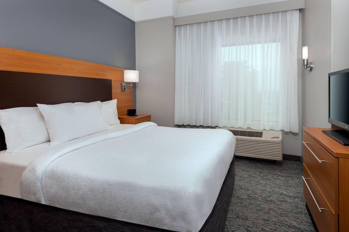 A hotel room with a large bed, a TV, a nightstand, a window with curtains, and a wall-mounted air conditioning unit.