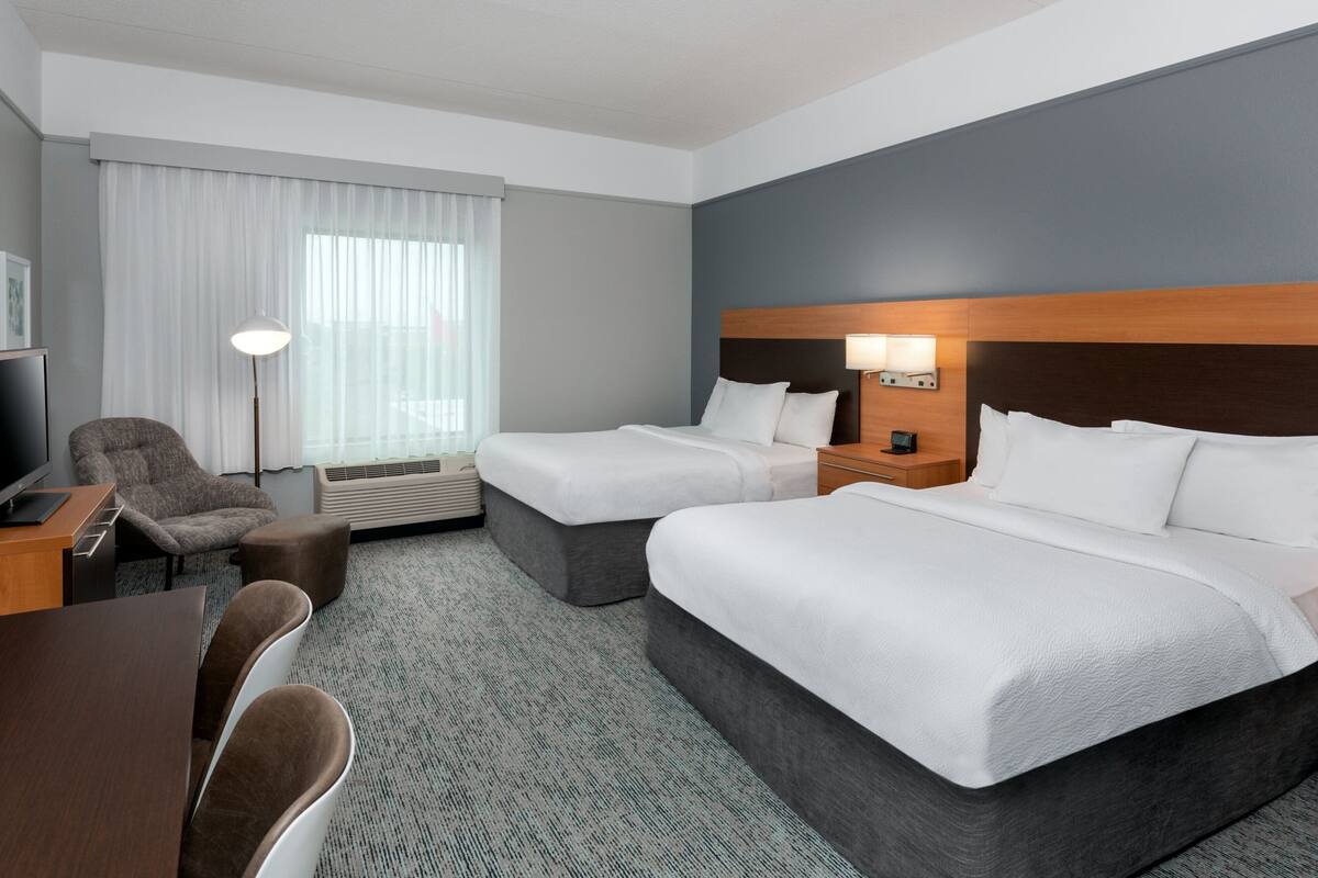 A hotel room with a large bed, a smaller bed, a desk with a chair, a TV, and a lamp.
