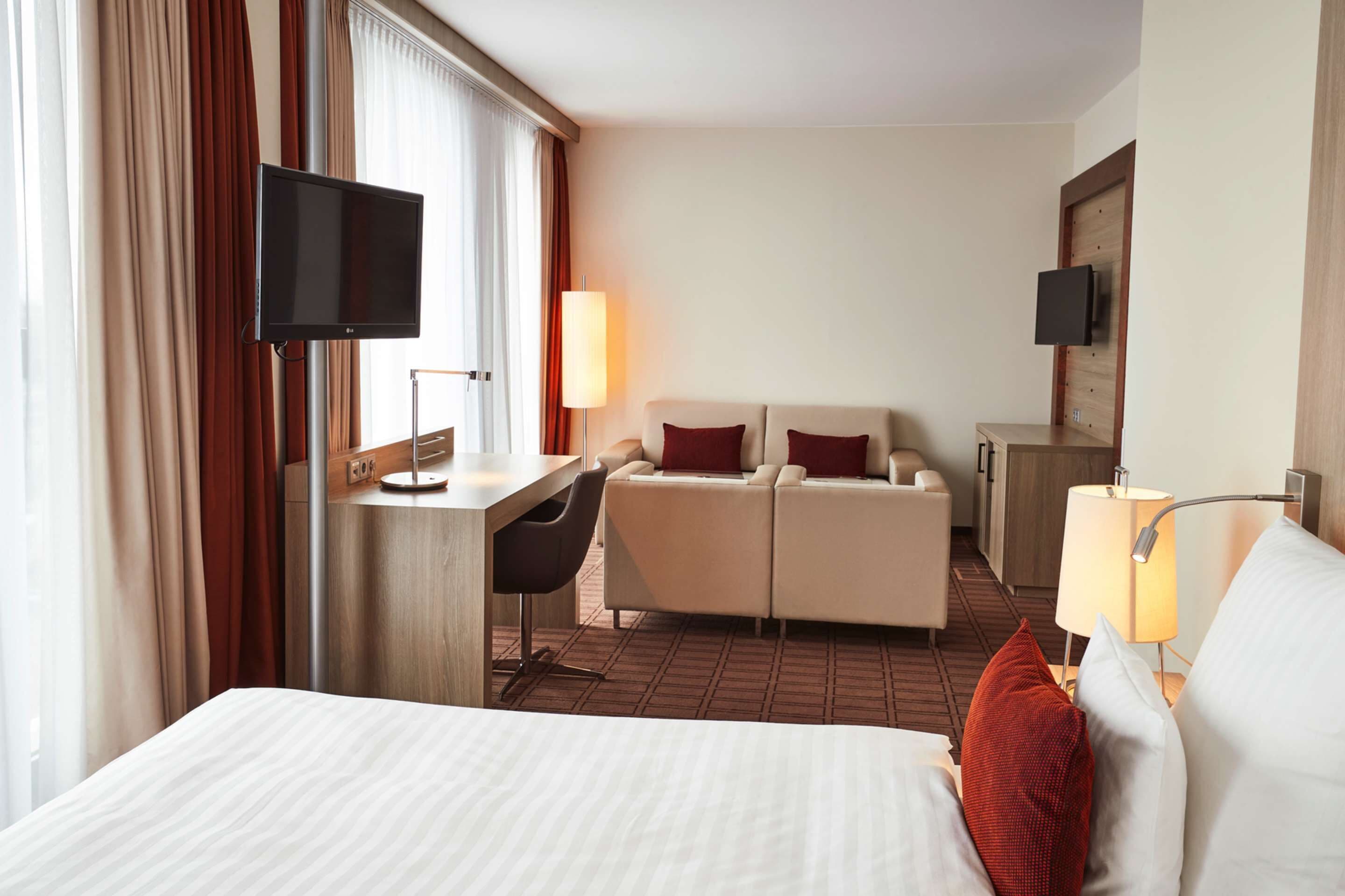 A hotel room with a bed, a desk, a chair, a sofa, two lamps, and a TV mounted on the wall.