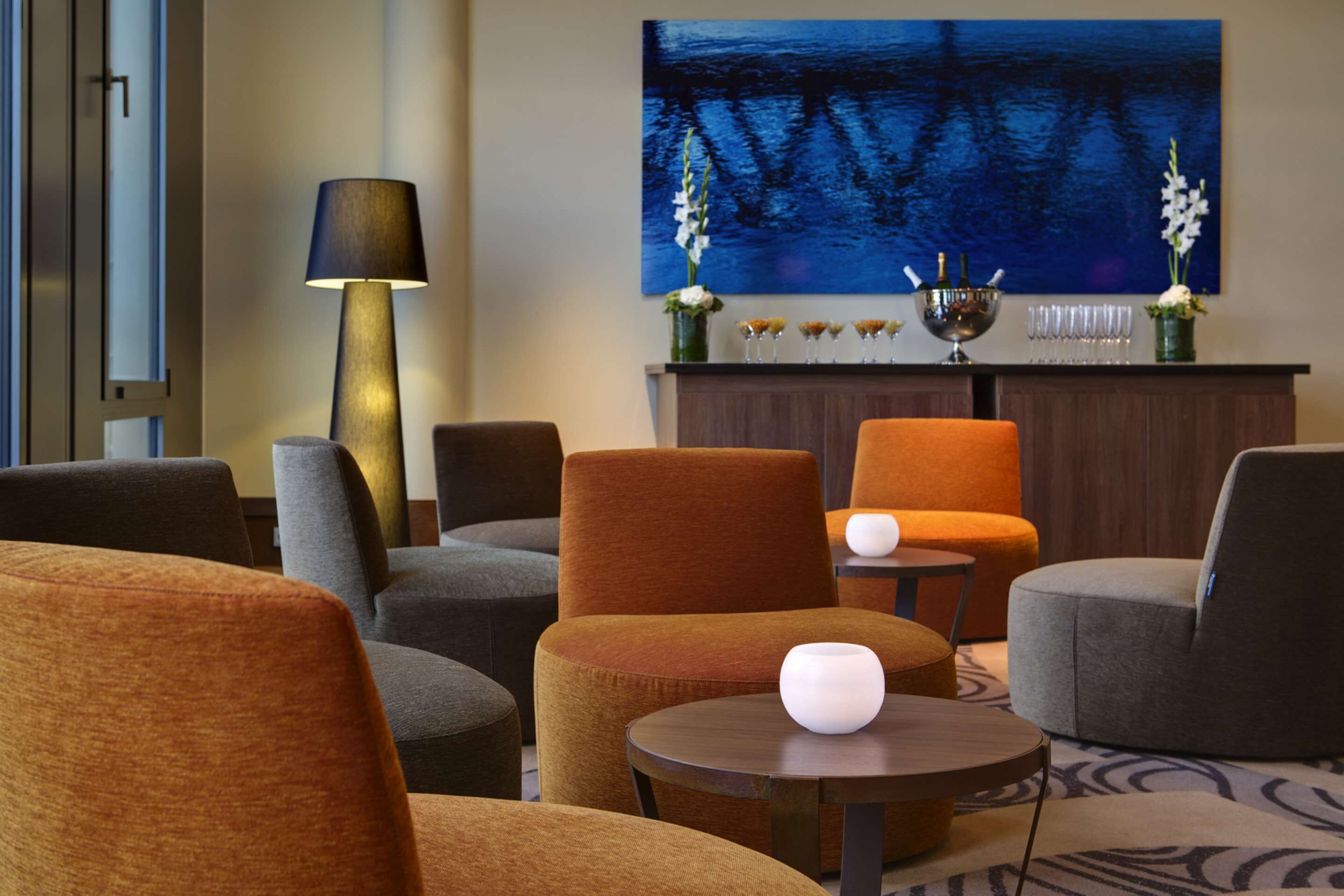 A modern lounge area with sofas and armchairs, a round table with a white vase, a floor lamp, and a large abstract painting on the wall.
