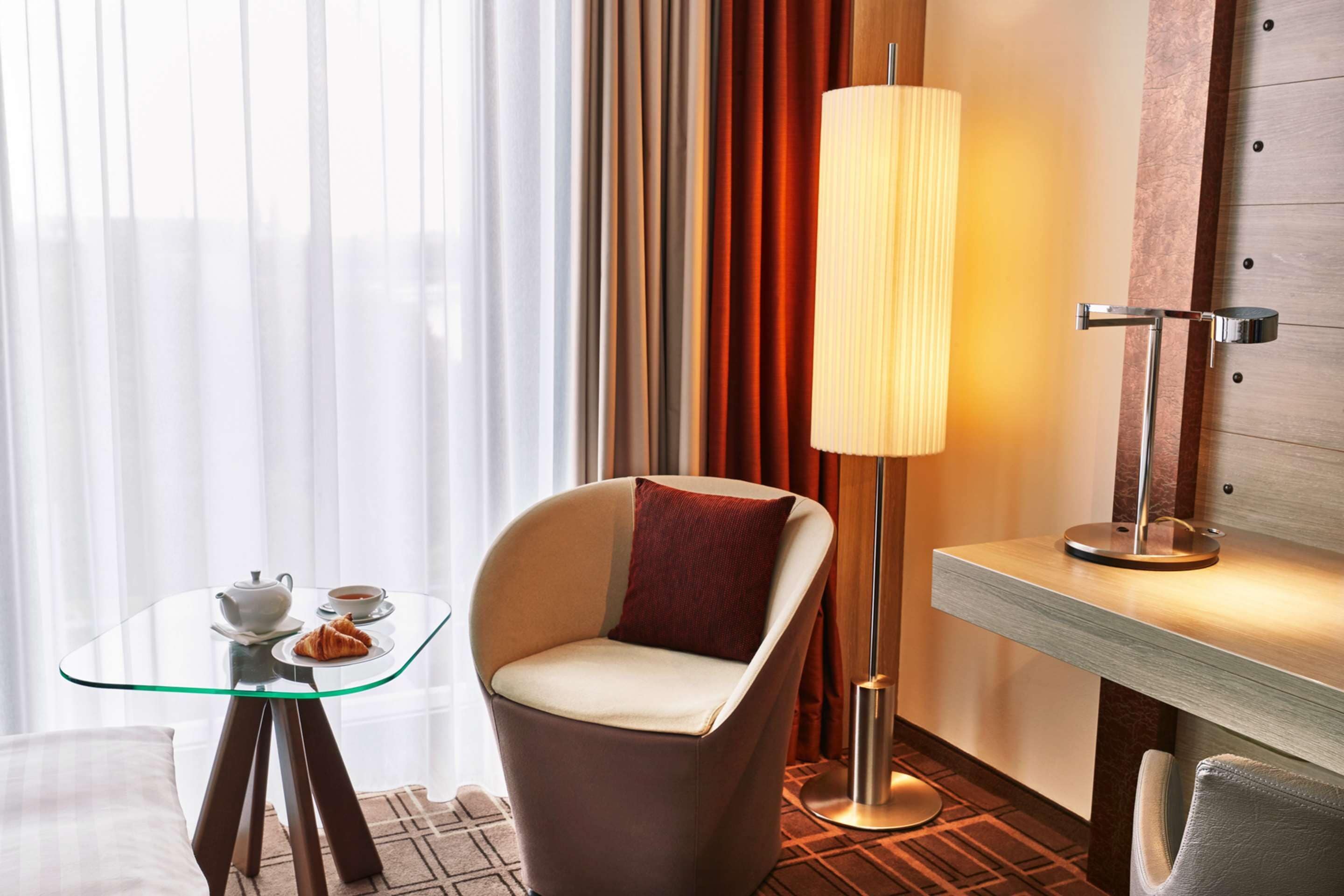 A modern hotel room with a glass table, a comfortable armchair, a standing lamp, and a view of curtains and a wooden wall.