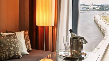 Habitación de hotel con sofá, almohadones decorativos, una mesa con una botella de champán y copas, y vistas a un río y edificios.