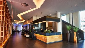 Un  área de recepción moderna de hotel, con mostrador de recepción, una escalera y un amplio ventanal.