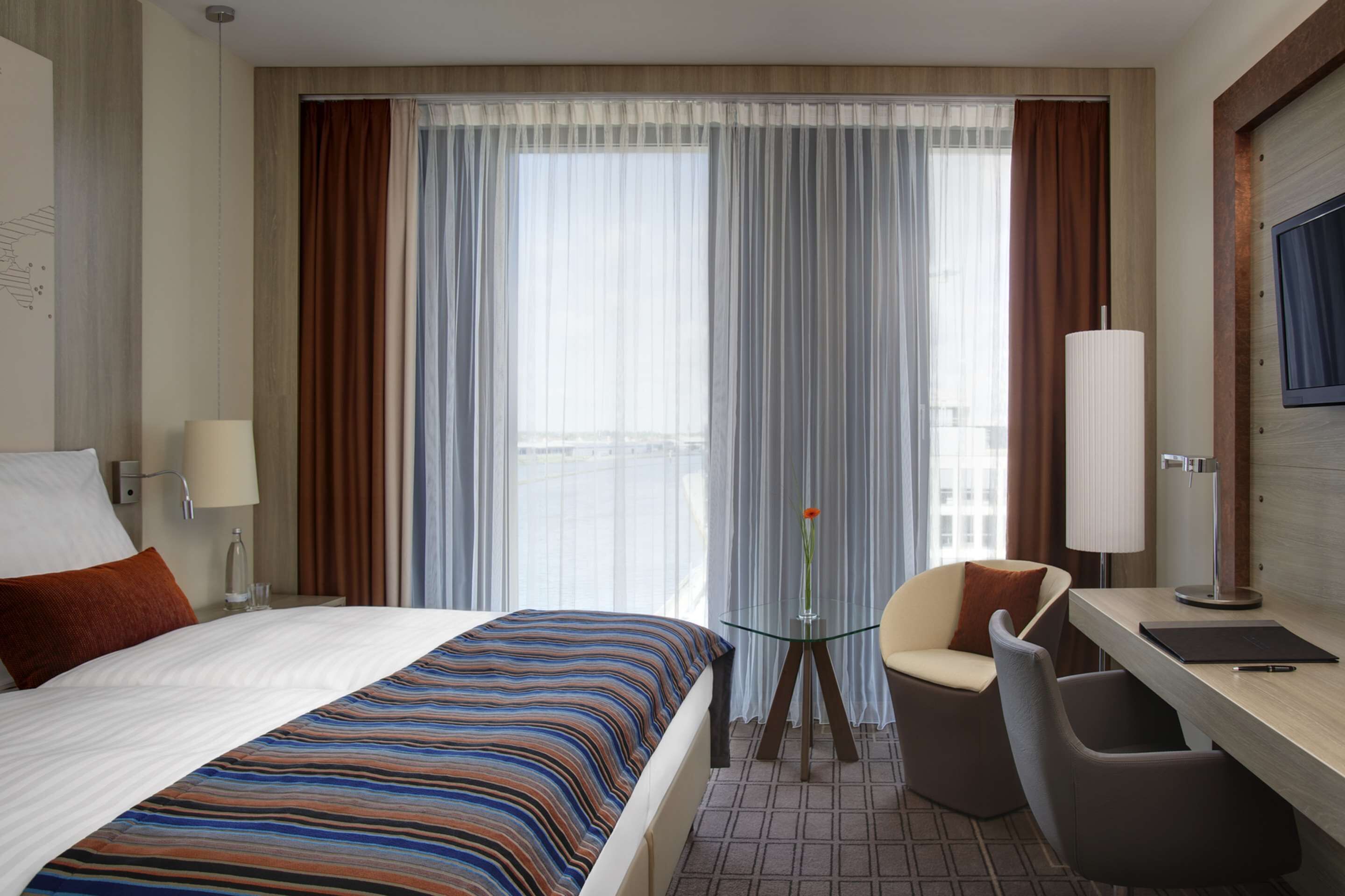 Una camera d'albergo con un letto, una scrivania, una sedia, una lampada e vista sulla città attraverso ampie finestre.