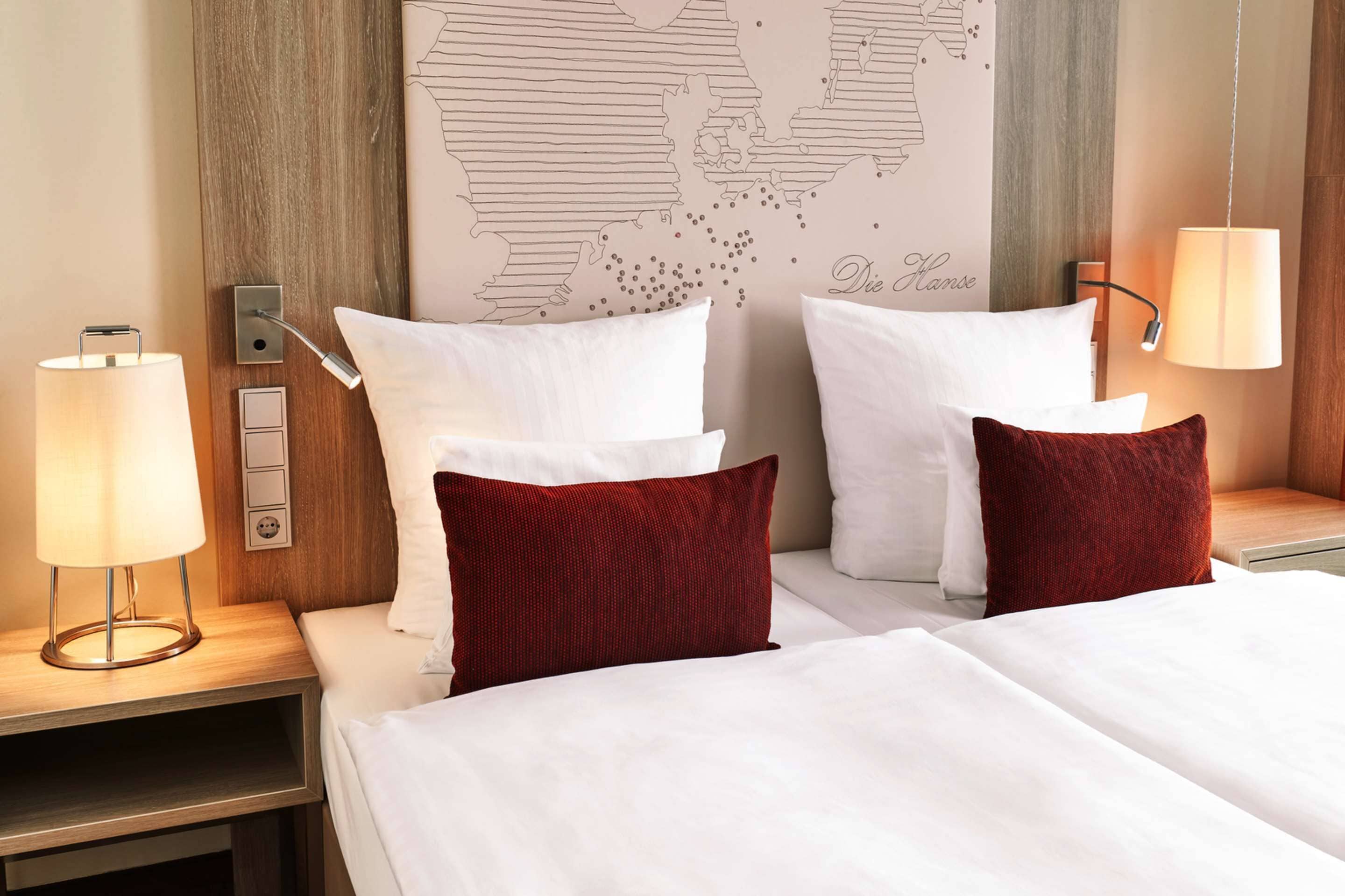 Habitación de hotel con dos camas, ropa de cama blanca, almohadas rojas, un mapa mundial en la pared y lámparas de noche.