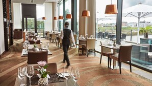 Un restaurante bien iluminado con mesas dispuestas para comer, amplias ventanas que ofrecen vista a la ciudad y un camarero caminando.