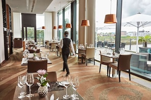 Un restaurante bien iluminado con mesas dispuestas para comer, amplias ventanas que ofrecen vista a la ciudad y un camarero caminando.