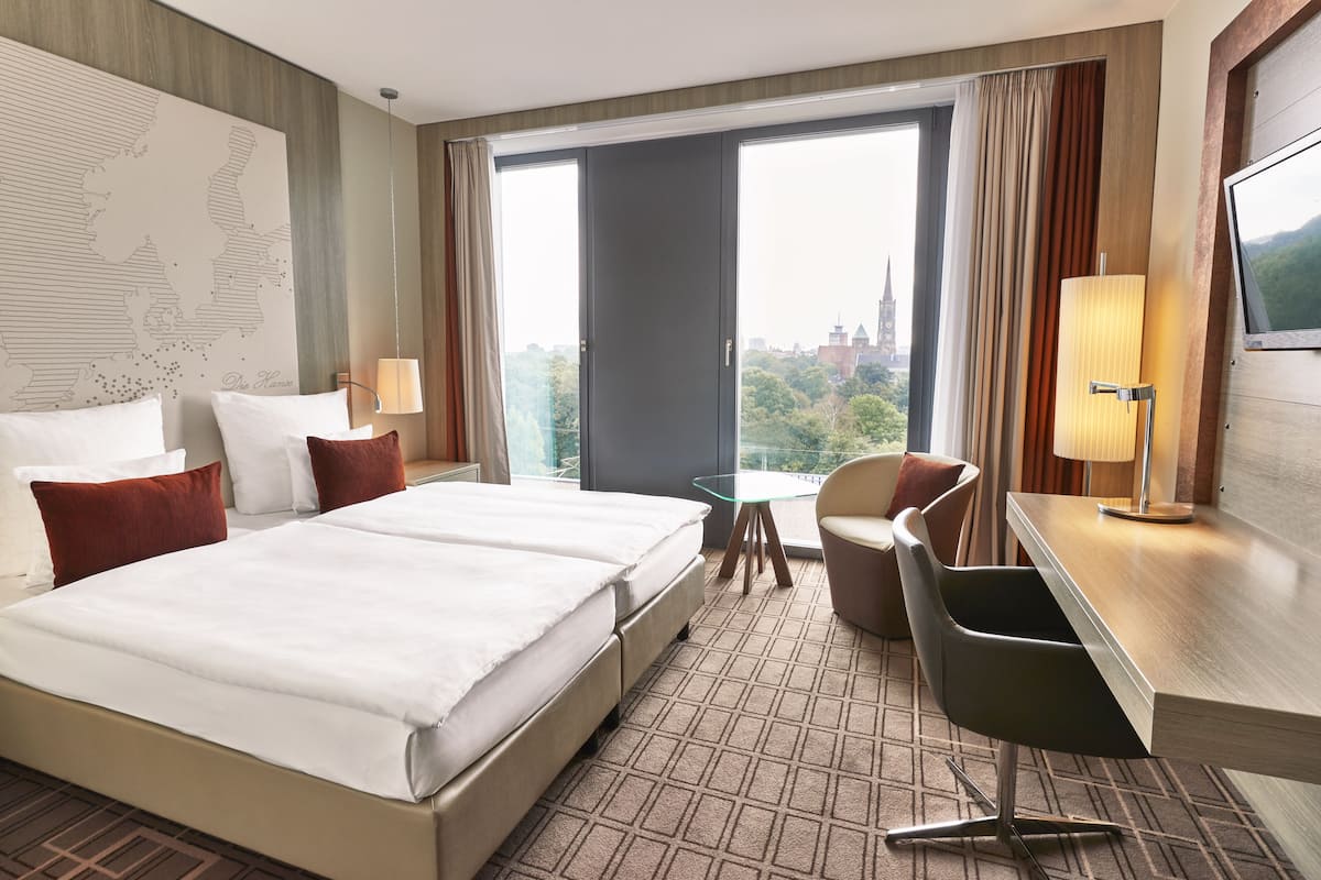 A hotel room with a large bed, a desk, a chair, a TV, and a window with a city view.