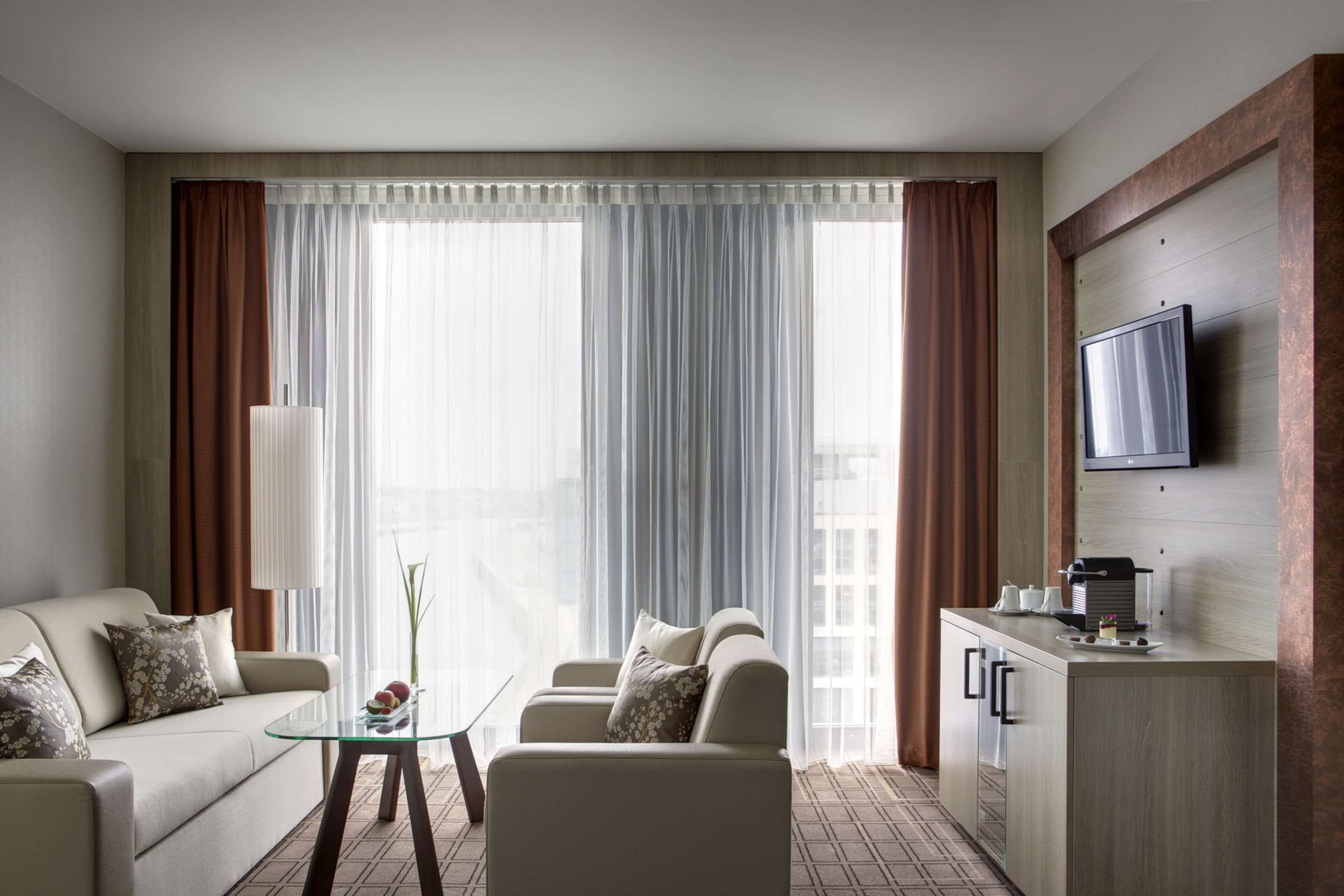 Een moderne hotelkamer met een bank, een glazen salontafel, een aan de muur bevestigde televisie en een klein kitchenette.