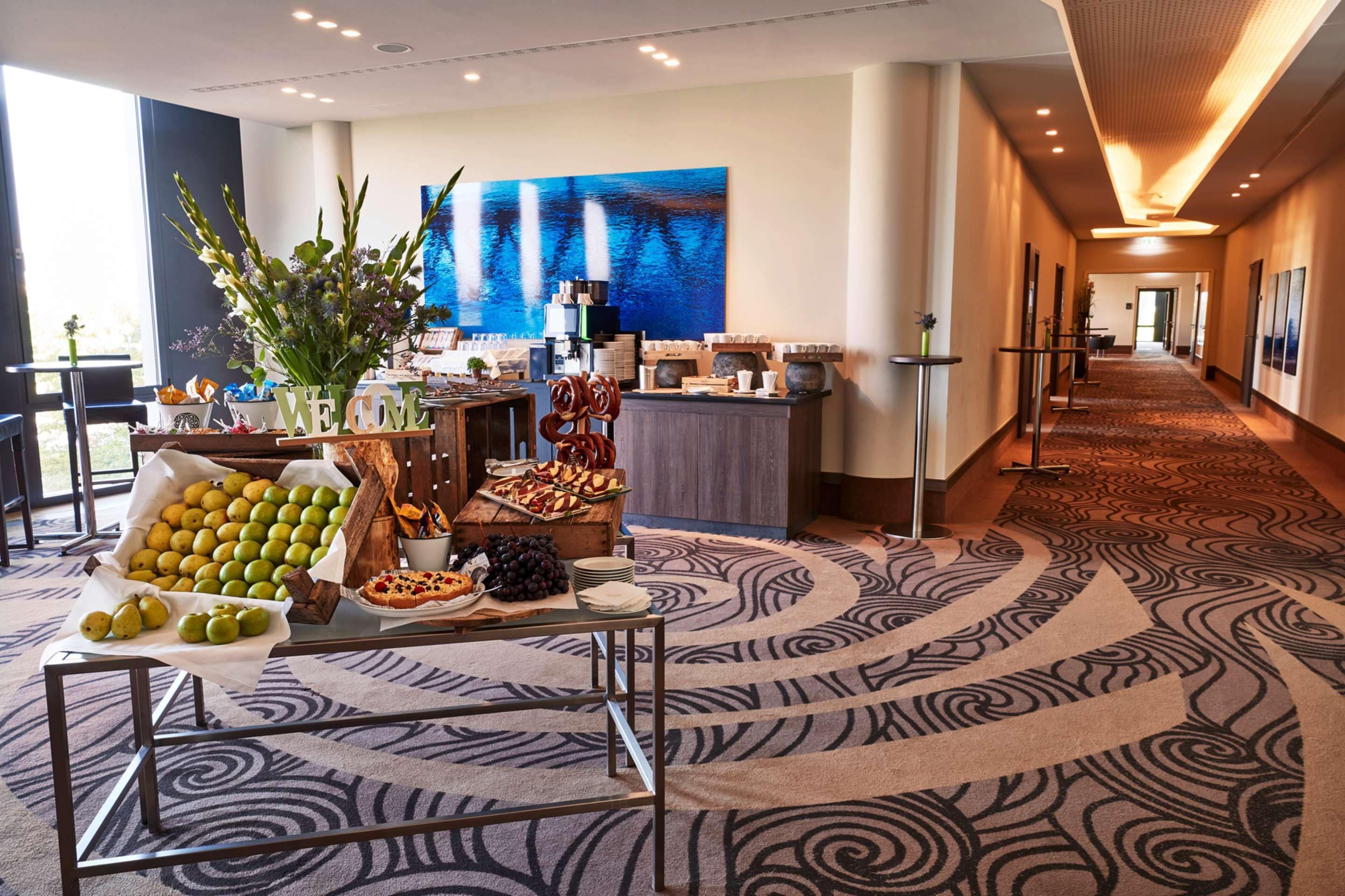 En hotellobby med reception, et stort abstrakt maleri, et buffetbord med frugt og bagværk, samt et tæppe med et mønster.
