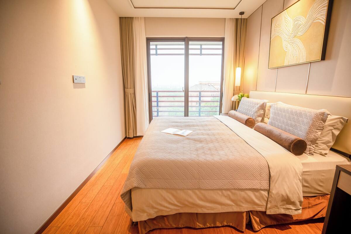 A hotel room with a large bed, a window with curtains, a wall-mounted artwork, and a bedside table with a lamp.
