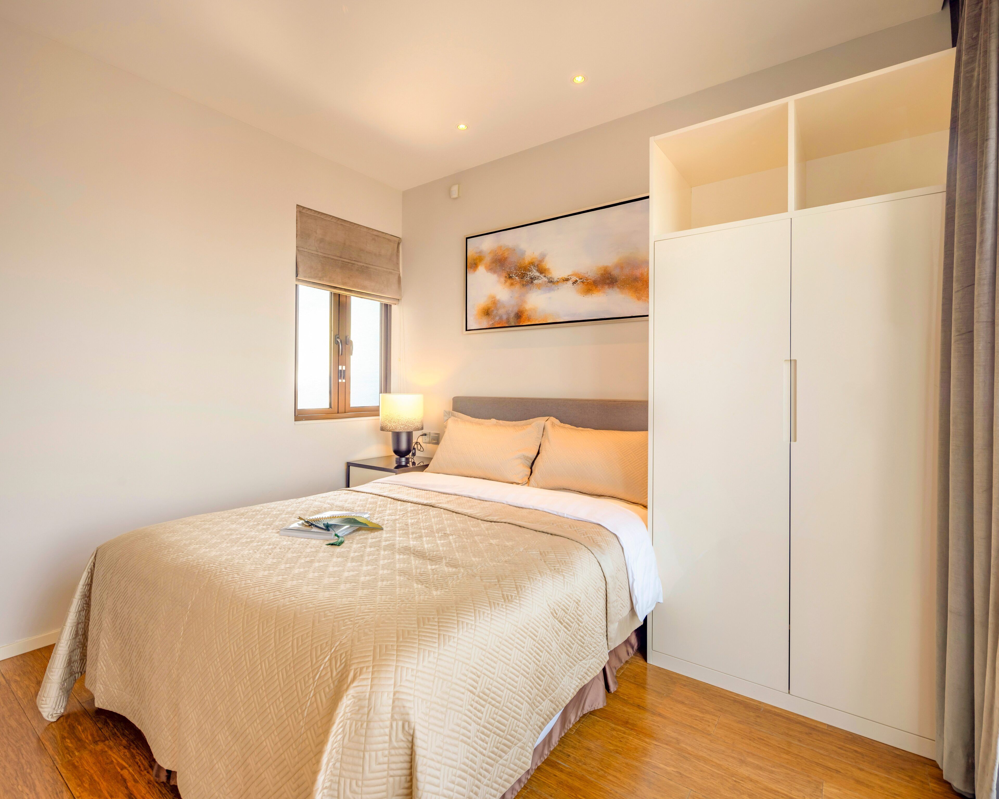 A bedroom with a bed, nightstand, lamp, window with blinds, artwork, and a wardrobe.