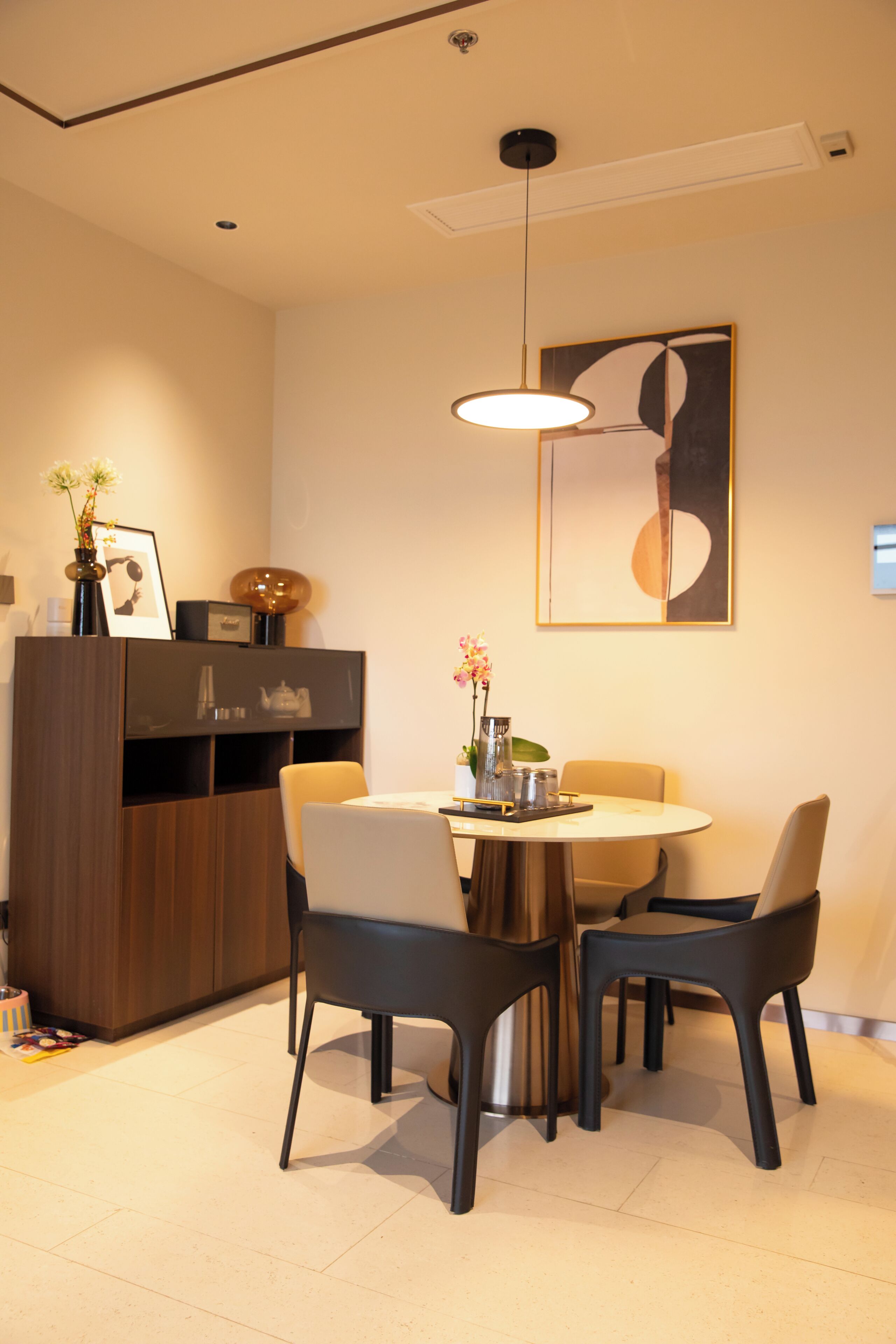 Un comedor con una mesa redonda, cuatro sillas, una lámpara colgante, un mueble y un cuadro abstracto enmarcado en la pared.